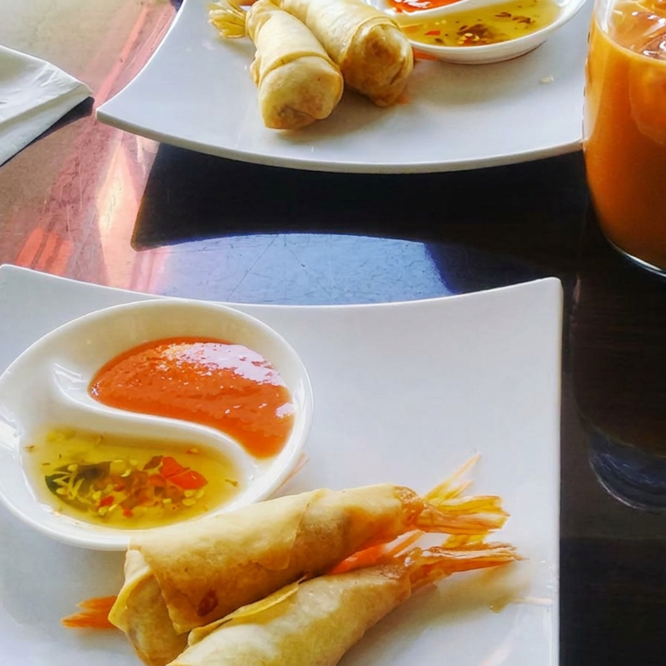 Crispy shrimp spring rolls with sweet chili dipping sauce, accompanied by iced orange drinks on a restaurant table.