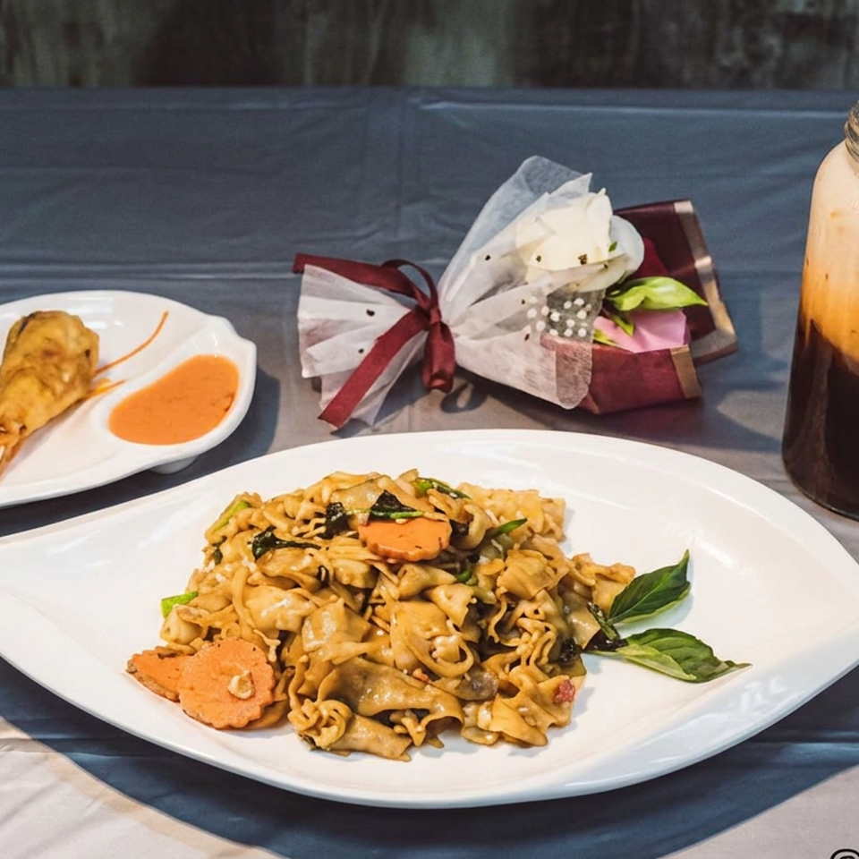 Stir-fried noodles with vegetables, fried shrimp and dipping sauce, a layered iced drink, and wrapped utensils on a table.
